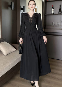 Full-length front view of a woman standing and wearing a black maxi dress. The gown features long bishop sleeves, a deep V-neckline with delicate black lace patchwork, a cinched, ruched waistline, and a long, flowing skirt. She carries a black quilted handbag.
