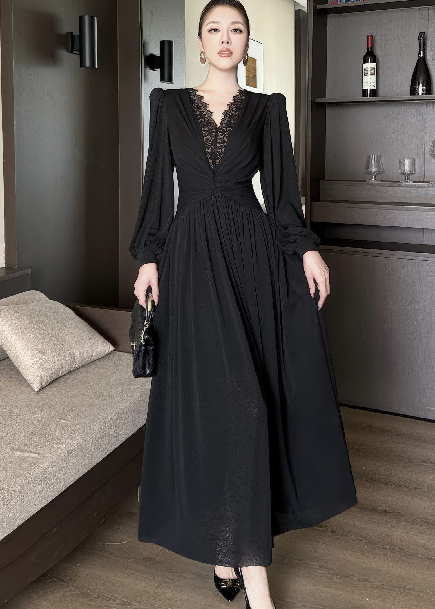Full-length front view of a woman standing and wearing a black maxi dress. The gown features long bishop sleeves, a deep V-neckline with delicate black lace patchwork, a cinched, ruched waistline, and a long, flowing skirt. She carries a black quilted handbag.
