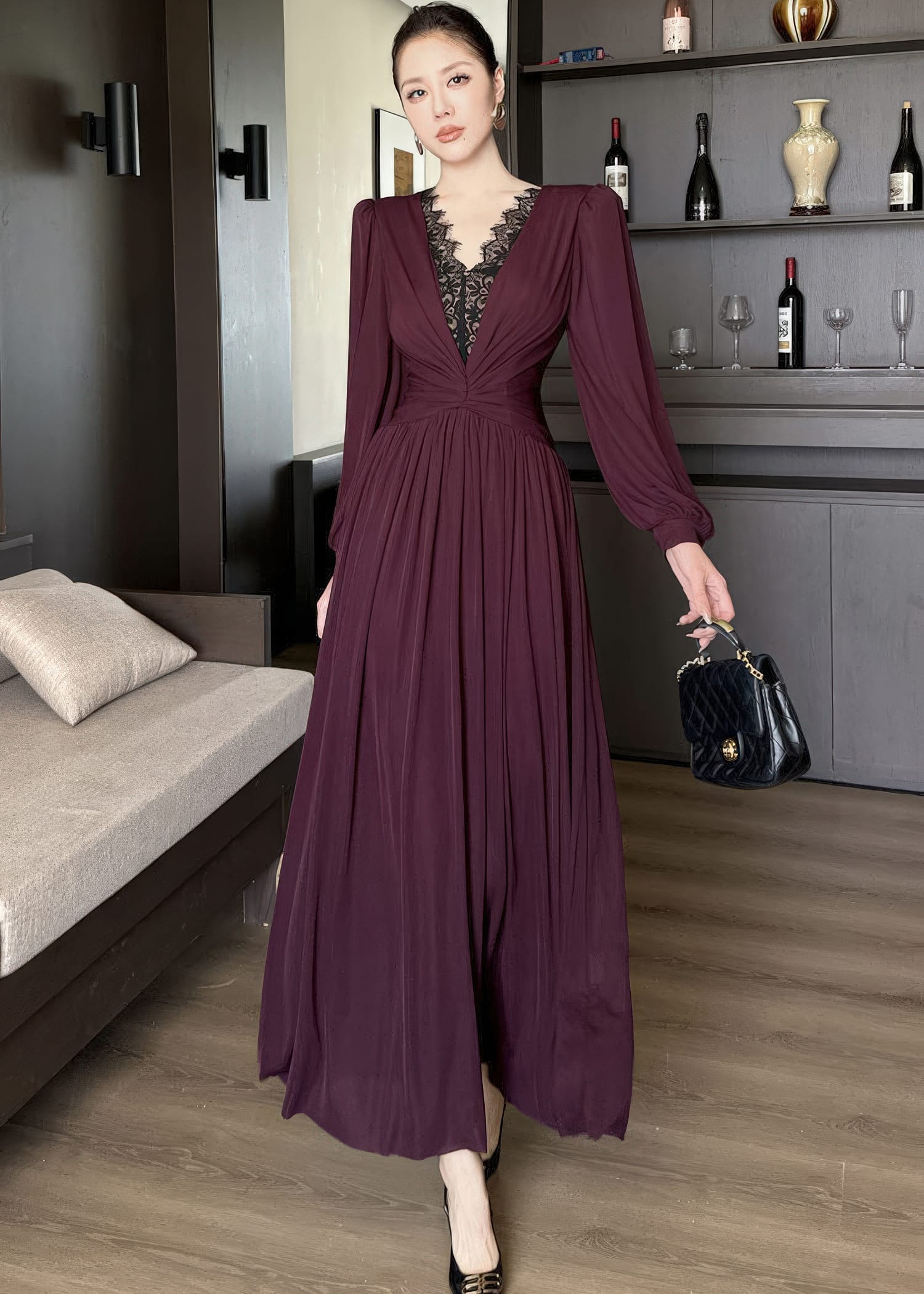 Full-length front view of a woman mid-stride, showcasing a burgundy maxi dress. The dress has long sleeves with cuffs, a prominent V-neck adorned with intricate black lace patchwork, a gathered bodice, and a full skirt. She holds a black quilted handbag in her left hand.