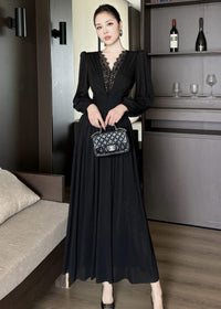 Full-length front view of a woman standing against a dark wood backdrop, wearing a black maxi dress. The design features a V-neckline framed with black lace patchwork, long puffed sleeves, a ruched fitted midsection, and a pleated floor-length skirt. She holds a black quilted handbag with both hands.