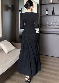 Full-length rear view of a woman standing in a black maxi dress, showing the back of the garment. The dress has a high back neckline, structured shoulders, a fitted waist, and a gathered skirt that flows to the floor. Her hair is tied in a bun, and she wears black heels.