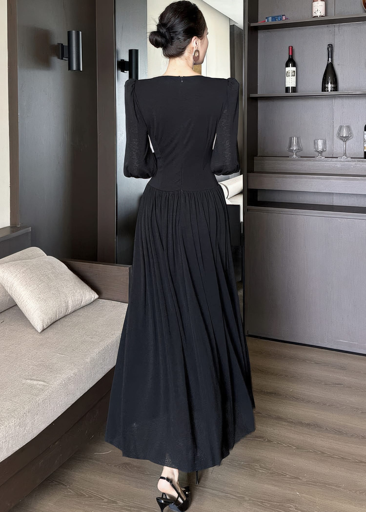 Full-length rear view of a woman standing in a black maxi dress, showing the back of the garment. The dress has a high back neckline, structured shoulders, a fitted waist, and a gathered skirt that flows to the floor. Her hair is tied in a bun, and she wears black heels.