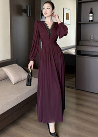 Full-length front view of a woman posing dynamically in a burgundy maxi dress. The dress features structured shoulders, long bishop sleeves, a deep V-neck with black lace patchwork details, a twisted ruched waist, and a flowing skirt. She holds a black quilted handbag.