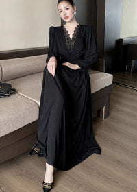 Full-length view of a woman sitting on a neutral-toned sofa, wearing a black maxi dress. The dress features a deep V-neckline with black lace patchwork, long sleeves, and a draped, floor-length skirt. She is wearing black heels with gold buckles.
