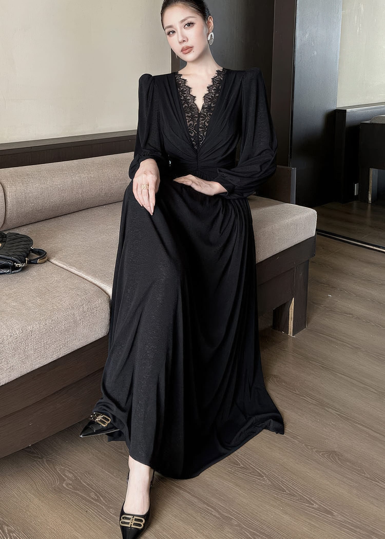 Full-length view of a woman sitting on a neutral-toned sofa, wearing a black maxi dress. The dress features a deep V-neckline with black lace patchwork, long sleeves, and a draped, floor-length skirt. She is wearing black heels with gold buckles.