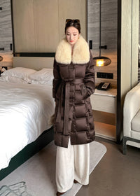 Full length front view of woman wearing brown quilted long down coat with cream fox fur collar and tied waist belt