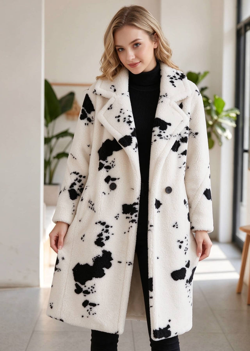 A woman wearing a long white faux-fur coat with black abstract splatter print, styled over a black turtleneck and black pants in a bright indoor setting.