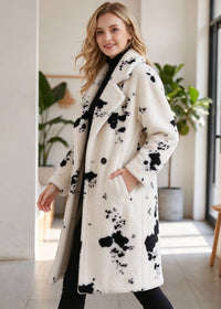 Front-facing walking pose of a woman modeling the oversized monochrome cow print notch lapel midi coat indoors. 