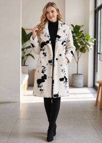 A woman modeling a cream-colored wool coat with black paint splatter patterns, worn over a black turtleneck and black pants in a bright indoor setting.