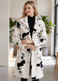 A woman wearing a long white faux-fur coat with black abstract splatter patterns, styled over a black turtleneck in a bright indoor setting.