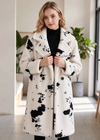 A woman wearing a long white faux-fur coat with black paint-splatter pattern, styled over a black turtleneck, standing in a bright indoor setting.