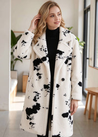 Front three-quarter portrait of an elegant woman touching her hair while wearing the cow print notch lapel faux shearling coat. 