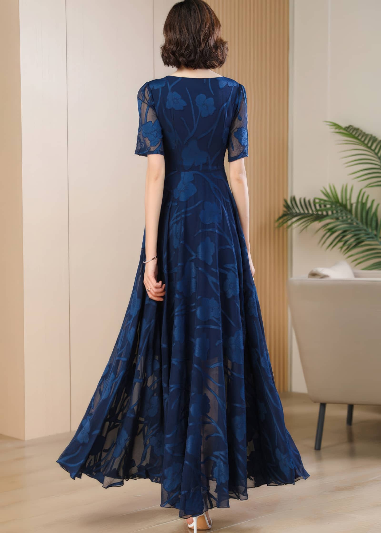 Rear view of a woman with short, wavy brown hair wearing a floor-length, short-sleeved navy blue maxi dress featuring a sheer floral burnout overlay.