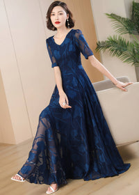 A woman in a navy blue floral burnout maxi dress leaning against a beige armchair, showcasing the flowy, floor-length skirt of the dress and white sandals.