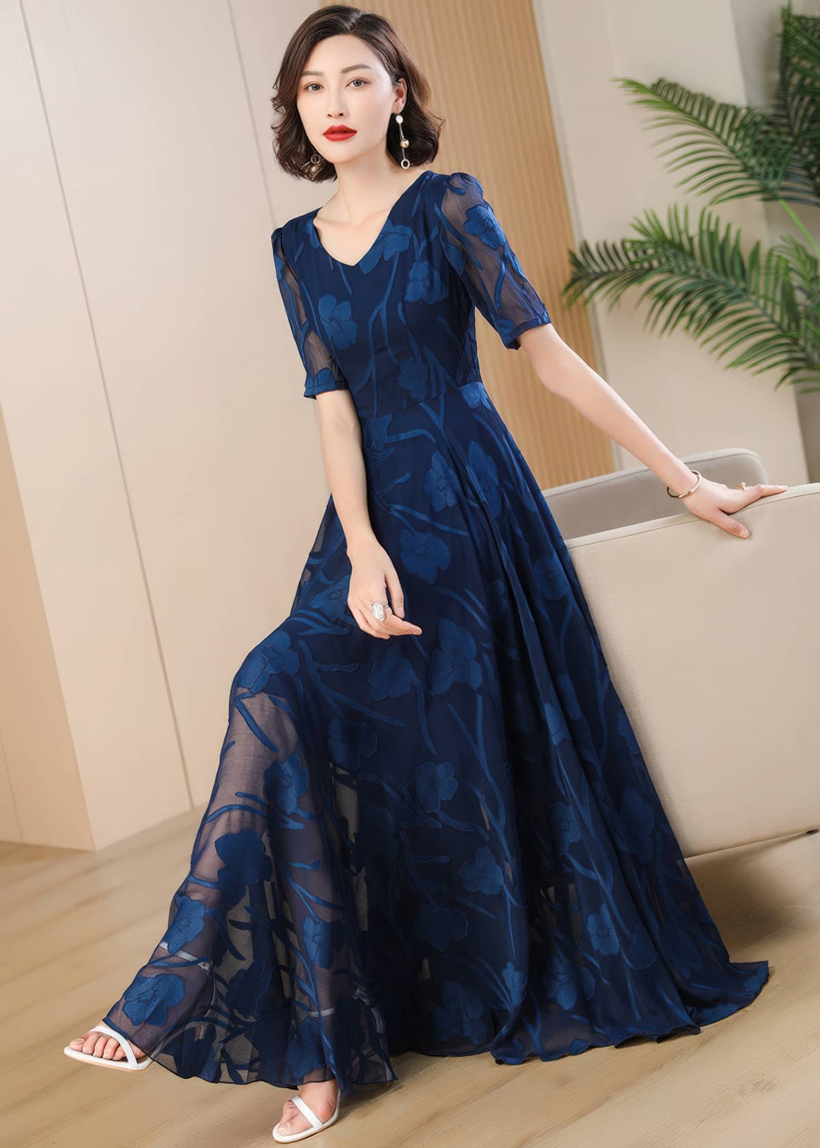 A woman in a navy blue floral burnout maxi dress leaning against a beige armchair, showcasing the flowy, floor-length skirt of the dress and white sandals.