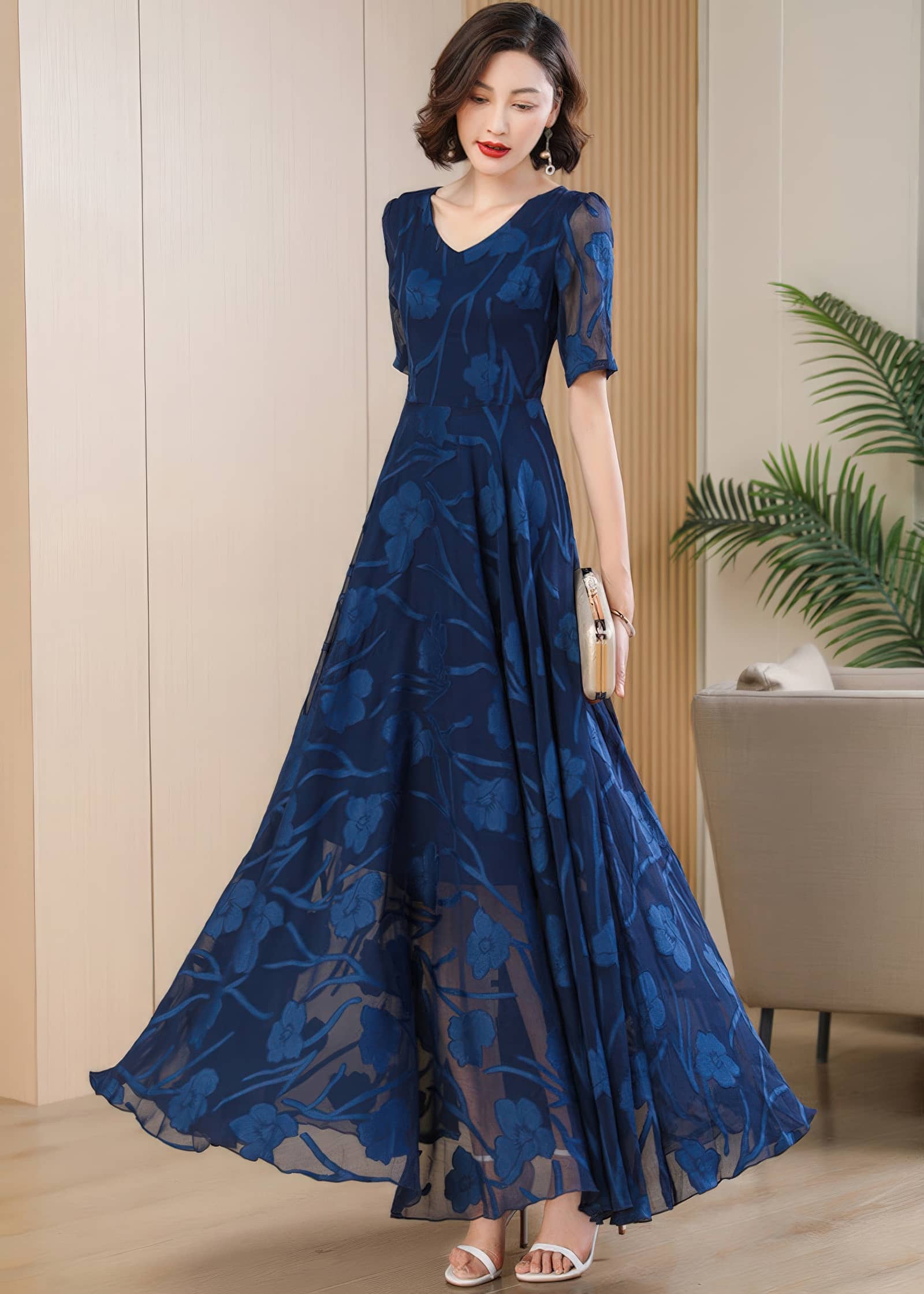 Three-quarter angle view of a woman posing in a navy blue floral burnout maxi dress while holding a gold metallic clutch purse.