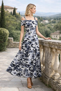 Profile view of a woman looking to the side while resting one hand on a stone balustrade, showcasing a black and white floral print off-the-shoulder maxi dress against a background of rolling hills.