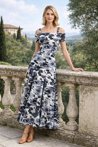 A woman leaning slightly against a decorative stone railing, displaying a full-length black and white floral print off-the-shoulder A-line dress.