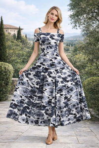 Front view of a blonde woman standing on an outdoor stone patio, holding out both sides of a black and white floral print off-the-shoulder A-line maxi dress.