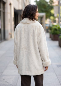 A woman viewed from behind wearing a fluffy cream faux fur coat on a city sidewalk.
