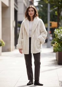 Full-body front view of a woman standing outdoors wearing an off-white faux fur coat, dark straight-leg pants, and ankle boots.   