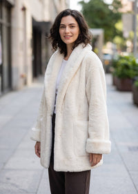 Three-quarter portrait of a smiling woman outdoors in a cozy off-white shawl-collar faux fur jacket.   