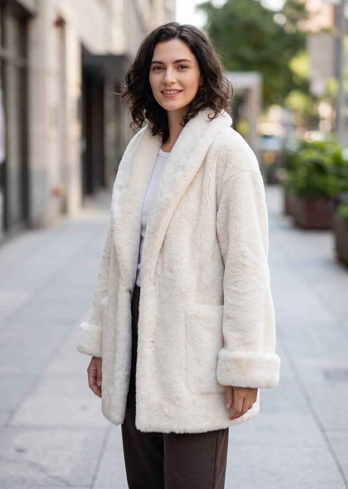 Three-quarter portrait of a smiling woman outdoors in a cozy off-white shawl-collar faux fur jacket.   