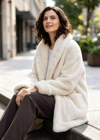 A woman sits on a stone curb outdoors wearing a plush cream faux-fur coat over a white top and dark brown pants, with a softly blurred city street in the background.