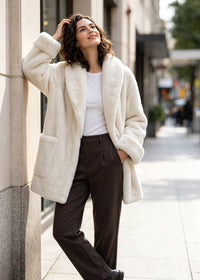 Full-length side pose of a smiling woman leaning against a wall wearing an open off-white faux fur coat with patch pockets.   