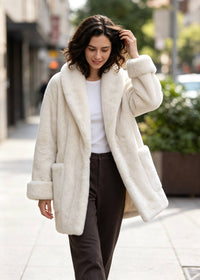 A woman walking on a city sidewalk wearing a plush cream-colored faux fur coat over a white top and dark brown pants.