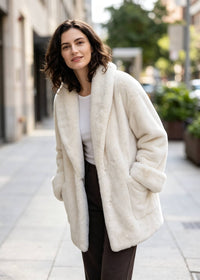 Three-quarter candid outdoor view of a woman with hands in pockets modeling an oversized off-white faux fur coat.   