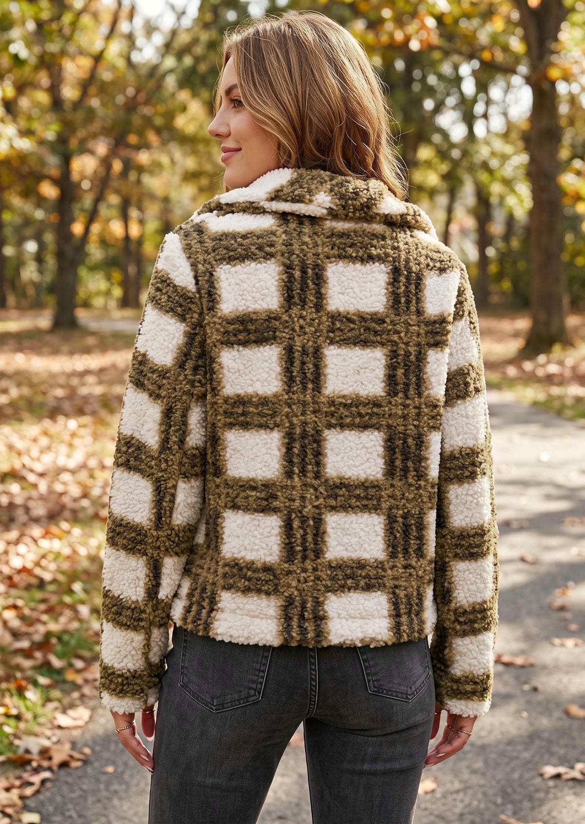 A woman wearing a fuzzy plaid hooded jacket in brown and cream, standing on an outdoor path surrounded by autumn trees.
