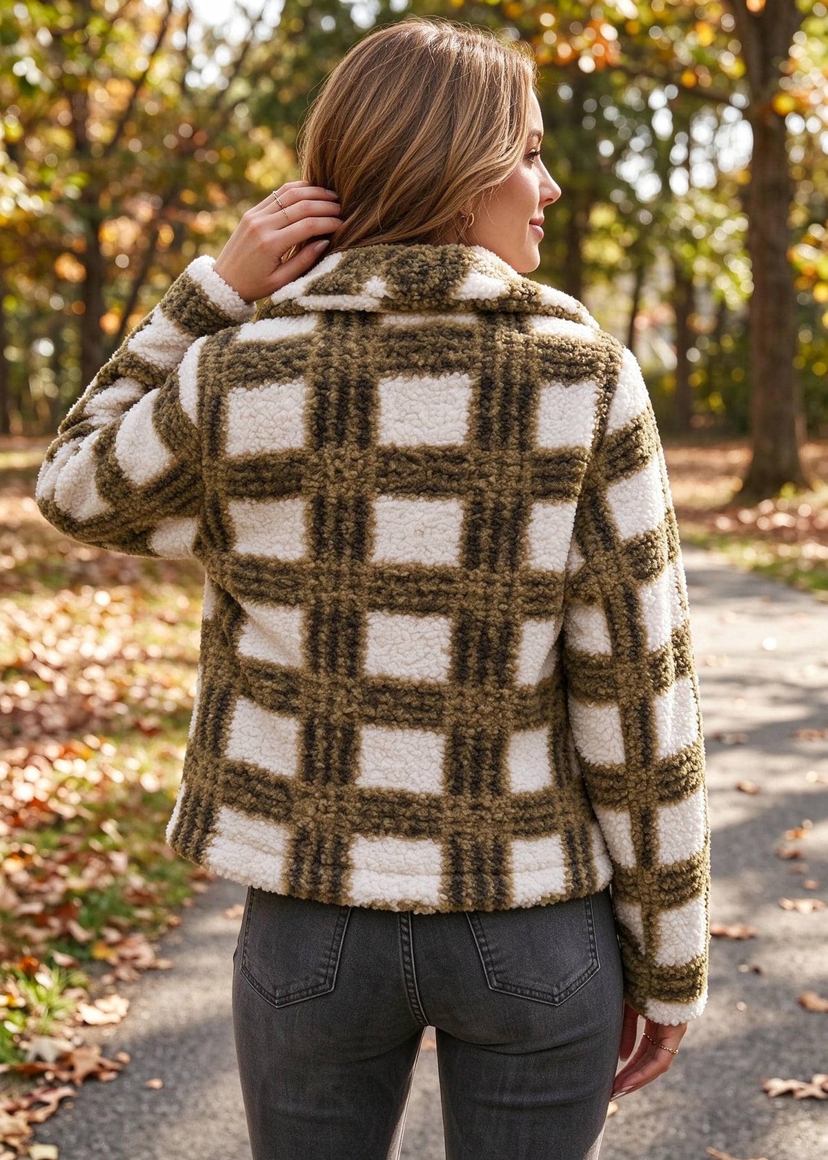 A woman viewed from behind wearing a cream and olive green plaid sherpa fleece jacket, standing on an outdoor path with fallen leaves and trees in the background.