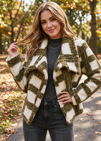 A woman wearing a textured, fuzzy open-front jacket with a green and cream geometric pattern, styled over a dark gray top and black jeans, standing on an outdoor path with autumn trees in the background.