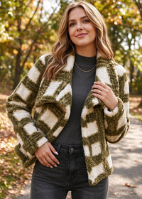 A woman wearing a fuzzy textured open-front jacket with a green and cream geometric pattern, styled with a dark gray top and black jeans, standing outdoors on a path with autumn trees in the background.
