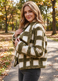 A woman wearing a fuzzy plaid cardigan in olive green and cream, standing on an outdoor path surrounded by autumn trees.