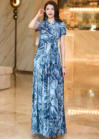 A portrait-style shot of a woman in a blue abstract print dress, lightly touching her hair. She wears long pearl drop earrings that complement the elegant, breezy style of the summer maxi dress.