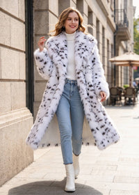 A woman walking on a city sidewalk wearing a long white faux fur coat with black spots, a white turtleneck, light blue jeans, and white ankle boots.