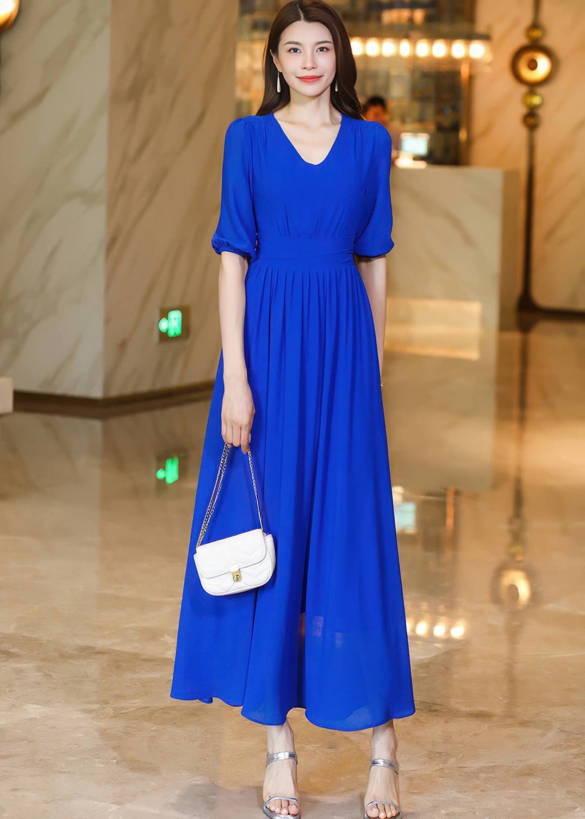 Front full-length view of a woman wearing a dusty pink V-neck chiffon midi dress with pleated detailing at the waist, styled with silver heels and a small white chain-strap bag.