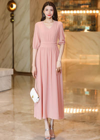 Front full-length shot of a woman in a dusty pink V-neck chiffon dress, styled with silver high-heeled sandals and a small white chain-strap purse.
