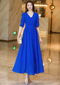A woman stands facing forward in a royal blue pleated chiffon midi dress with silver heels and a white clutch bag.