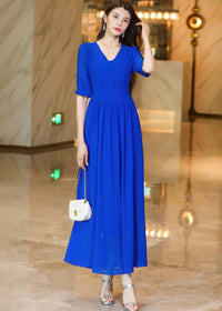 Frontal shot of a woman in a royal blue chiffon midi dress, holding a white purse and posing with one hand near her hair.