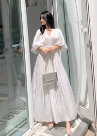 A full-length shot of a woman in a flowing white pleated dress, posing with both hands on a silver chain-strap purse. The dress features a cinched waist with beaded detailing and voluminous puff sleeves.