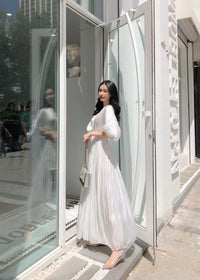 A side-profile view of a woman standing outside a white building wearing an elegant white pleated gown. The fabric has a subtle metallic sheen, and she is wearing clear, iridescent flat shoes.