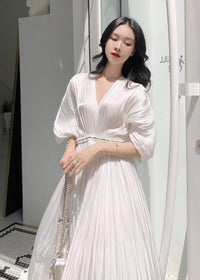 A medium close-up of a woman wearing a white pleated V-neck dress. The image highlights the texture of the vertical pleats, the puff sleeve design, and her delicate jewelry, including a large silver ring.