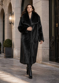 Front view of woman holding the front lapel of a plush black wide-collar faux fur coat paired with knee-high leather boots