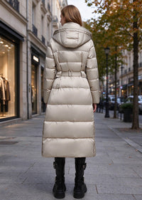 A woman viewed from behind wearing a long, puffer-style hooded coat in a light champagne beige color, standing on a city sidewalk.