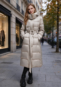 A woman wearing a long, shiny puffer coat in a light beige or champagne color, standing on a city sidewalk. The coat has a high collar, front zipper, belt, and front pockets.