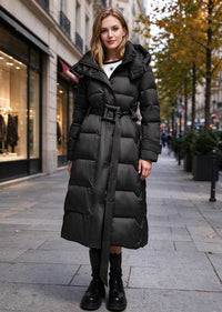 A woman stands on a city sidewalk wearing a long black puffer coat with a belted waist, black knee-high socks, and black boots. The background shows a blurred urban street scene with storefronts and trees.