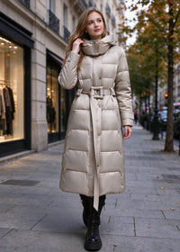A woman wearing a long, light beige puffer coat with a belted waist and high collar, walking on a city sidewalk.